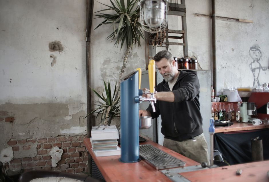 Brutal male in casual wear standing at counter in grungy garage and pouring water from dispenser while relaxing during work